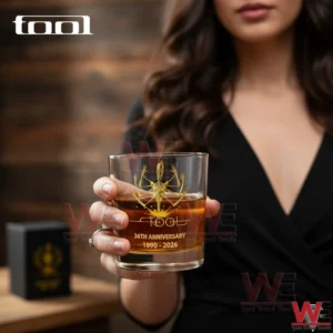 Tool Band Alex Grey Tool figure 1990-2026 Glass 5 A close view of a TOOL anniversary whiskey glass held by a woman showcasing the gold emblem and celebratory text against a soft background!