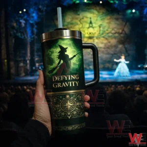 Hand holding a green Defying Gravity witch tumbler with handle and straw inside a theater with a blurred stage in the background.