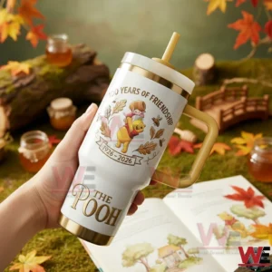 Winnie the Pooh 100th Anniversary A Century of Friendship Tumbler 3 Hand holding a white and gold The Pooh 100 Years Of Friendship insulated tumbler with handle and straw featuring Pooh artwork and anniversary dates with honey jars autumn leaves and an open book in the background!