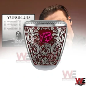 Side view of silver custom Yungblud ring with red ornate filigree panel and pink YB monogram against a blurred poster background!