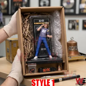 A high quality product photograph shows an open brown cardboard box held by two gloved hands revealing a nested AC DC Bon Scott figure with amplifier! The figure is protected by form fitting brown paper shreds! Below the figure are accessories including a pouch with picks a guitar strap with the AC DC logo and a miniature bell! The box rests on a wooden desk with a utility knife and pencils and framed awards are visible in the blurry background! The lighting is clear and even!