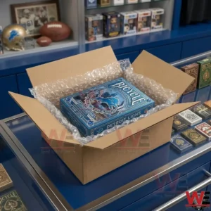 The open cardboard box, filled with clear plastic bubble wrap, holds the detailed blue Bicycle Detroit Lions 95th Anniversary Commemorative card deck, resting diagonally, with its face and top visible. The setting is a counter with multiple other card decks and sports memorabilia on display and on shelves in the background.