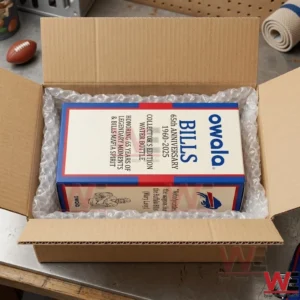 An open cardboard box contains a white Owala collector box for a Buffalo Bills anniversary water bottle. The item is protected by clear bubble wrap and sits on a metal table with a small football nearby.