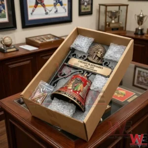 An overhead view of the Chicago Blackhawks anniversary bell packed in a shipping box! The box contains foam padding bubble wrap and a bag of screws! It is placed on a glass counter in a sports memorabilia room!