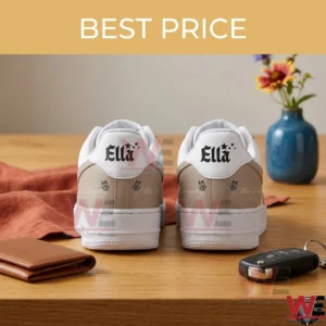 A rear view photograph of two white and tan low top sneakers sitting on a wooden table! On each shoe the white heel tab features the name Ella and small star graphics in black! Two black butterfly outlines are printed on the tan leather heel section of both shoes! Surrounding objects include a brown wallet a car key dried flowers in a blue vase and an orange textile!