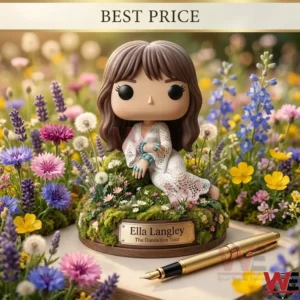 A vinyl collectible figure of a woman in a white dress sitting on a flowery moss mound! The figure has brown hair and turquoise accessories! A gold nameplate on the wooden base identifies her as Ella Langley! The scene is surrounded by colorful wildflowers including dandelions and lavender! A gold pen is on the table next to the figure!