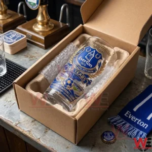 An Everton anniversary glass is packed in a cardboard box with protective inserts on a bar counter. A blue team scarf and a bottle cap are nearby on the counter surface.