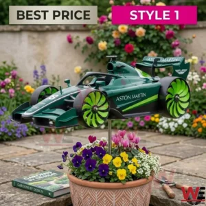 A close-up photograph of a green Aston Martin F1 model car on a black stand in a pot of spring flowers within a garden setting. The car has green, fan-shaped wheels.