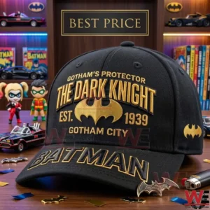 A black cap with gold Batman embroidery sits on a table! The design includes the words The Dark Knight and Est 1939 with a bat logo! Batman figurines and comic books are in the blurred background!