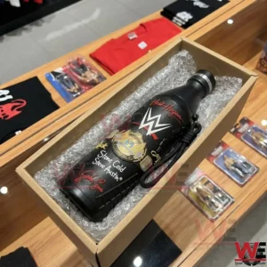 A black WWE water bottle is packed inside a cardboard box with protective bubble wrap. The box is on a wooden shop counter with wrestling shirts and toys visible in the background.