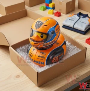 A McLaren racing rubber duck is packaged inside a cardboard box with bubble wrap. The box is on a wooden table with team merchandise and building blocks nearby.