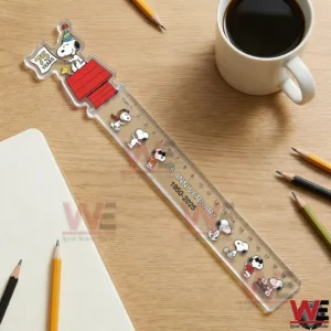 Clear Snoopy 75th anniversary ruler on a wooden desk beside a coffee mug, notebook, and pencils!