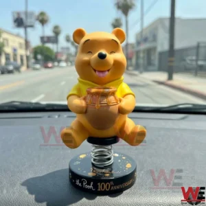 Yellow bear bobblehead toy on a car dashboard with a sunny street background.
