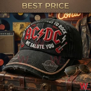 A close-up photograph shows a black washed denim baseball cap with an embroidered AC/DC logo, a lightning bolt, and rock band text, resting on a worn leather trunk with sunglasses. The background features rock memorabilia.