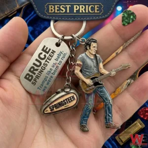 A close up photograph of a hand holding a large metal keychain! Three charms hang from the main ring a detailed figure of Springsteen a metal tag with his name and a song lyric and a replica of a classic motorcycle fuel tank! Other music accessories like picks and a harmonica are visible in the background!