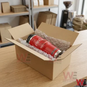 An open cardboard box lined with bubble wrap contains the red Coca-Cola insulated tumbler. The box is on a wooden counter in a shipping and logistics environment with shelves and coffee equipment.