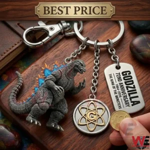 A close up photograph of a Godzilla anniversary keychain with three charms on a wooden desk! The charms include a Godzilla figure with glowing fins an atomic symbol medallion and an engraved metal tag! A hand holds a coin next to the keychain!