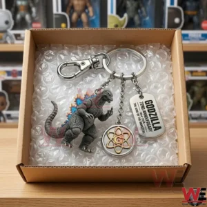 An overhead view of a Godzilla anniversary keychain inside a shipping box with bubble wrap! The three charms are neatly arranged! The background shows a room with shelves full of toy collectibles!