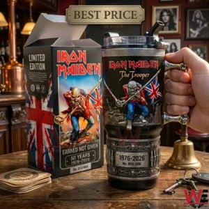 A close up photograph of an Iron Maiden themed tumbler and its packaging box on a wooden table! The tumbler has a unique bone handle and detailed carvings! A bell and keys are placed nearby!