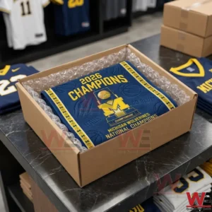 The image shows a square cardboard box sitting on a dark counter! Inside the box a blue towel with yellow championship branding is visible! The towel is surrounded by clear bubble wrap for protection! The background reveals a retail environment with folded jerseys and other shipping boxes stacked on shelves! The lighting is bright and clear!