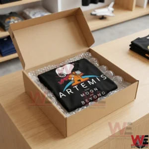 A folded black Artemis mission t shirt inside a cardboard shipping box on a wooden desk!