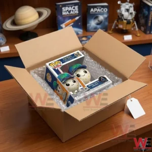 An open cardboard shipping box lined with bubble wrap, containing the NASA Moon Mascot Rise collectible box from image_1.png! Through the clear window, the Moon figure with its cap and rockets is visible! A small blank tag is attached to the shipping box!