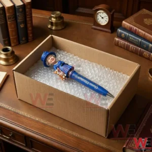 A blue commemorative pen with a small figurine is packed in a cardboard box with bubble wrap! The package sits on a wooden table among vintage books and a clock!