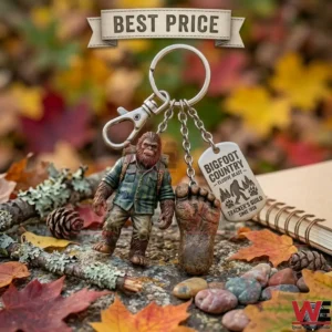 Sasquatch themed metal keychain with a Bigfoot figurine and a tracking tag on a forest floor.